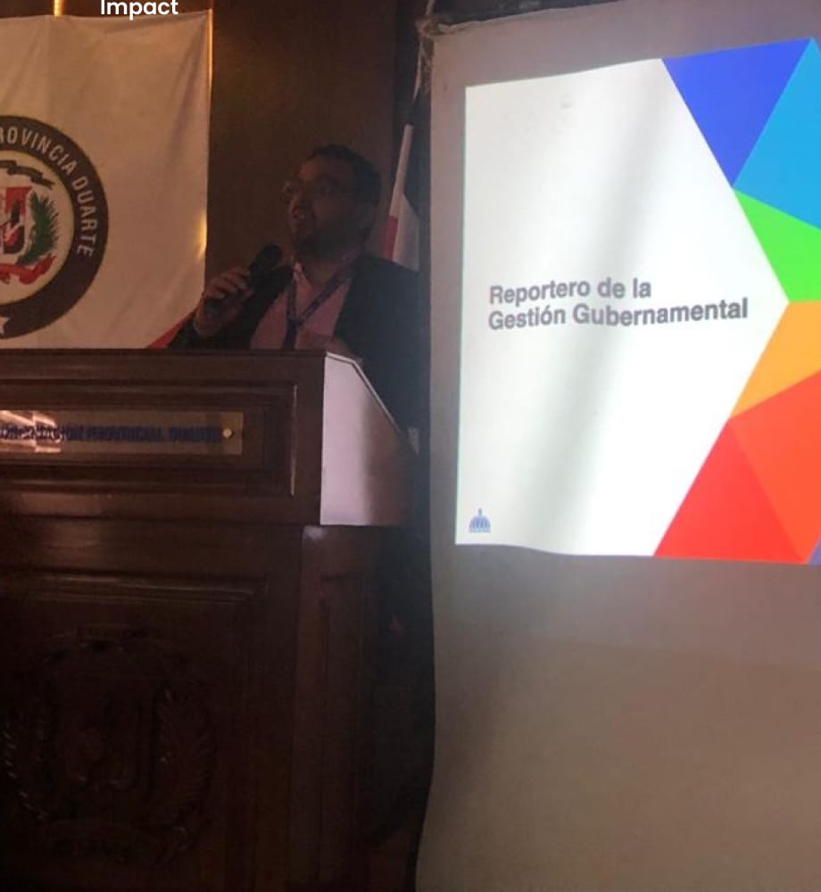 Fran Bencosme presenting Reportero at a lectern in Provincia Duarte, the project slide on the screen beside him